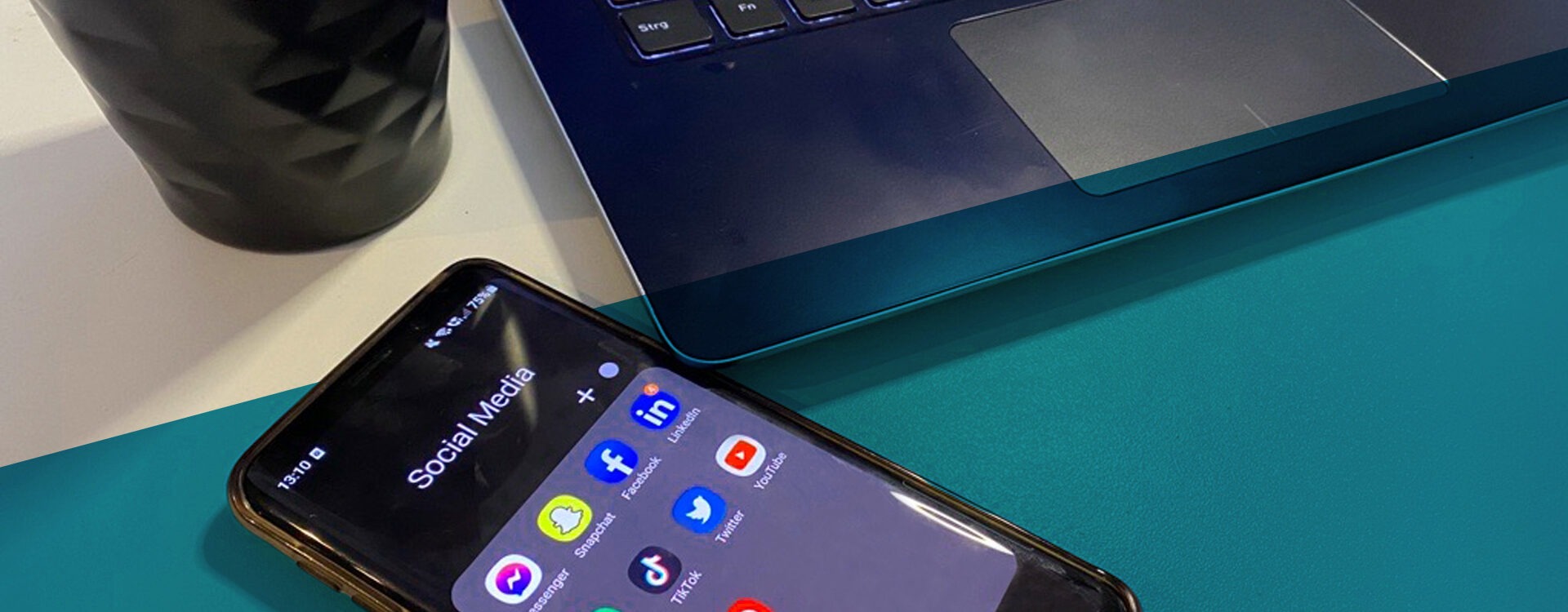 Handy liegt neben Laptop und Kaffeebecher auf dem Schreibtisch. Auf dem Handy sind diverse Icons sozialer Netzwerke zu sehen