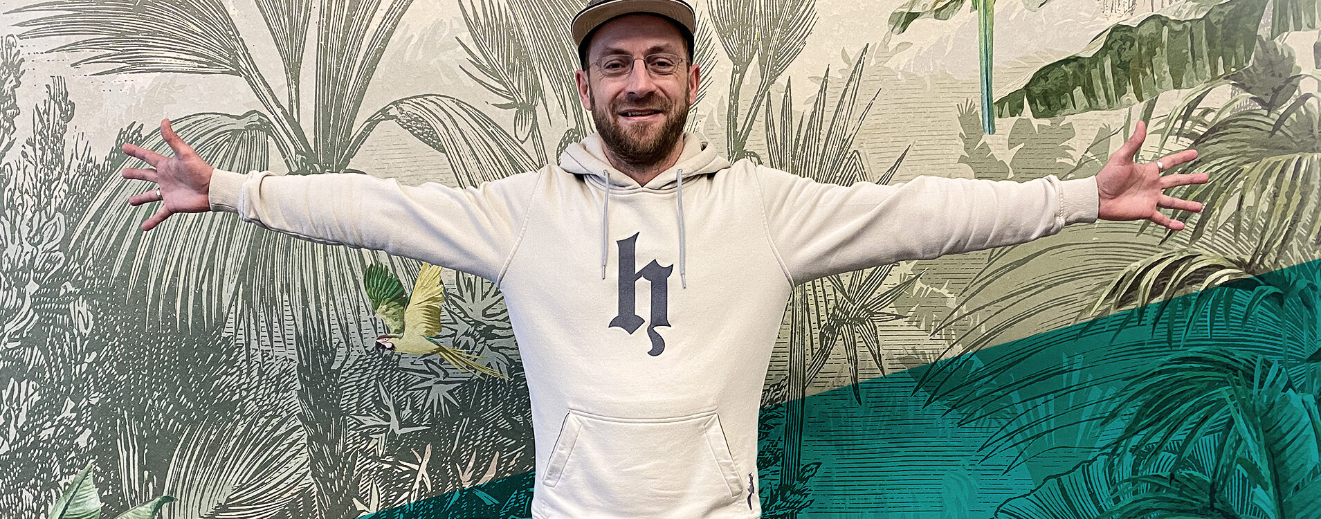 Mann mit beigem Oberteil und Cap steht mit ausgebreiteten Armen vor einer Tapete im Dschungel-Design