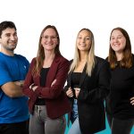 Vier Personen, drei Frauen und ein Mann, stehen vor einem blau-weißen Hintergrund und lächeln in die Kamera