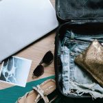 Utensilien für den Urlaub (Laptop, Koffer, Sonnenbrille) ausgebreitet auf einem Tisch, von oben fotografiert