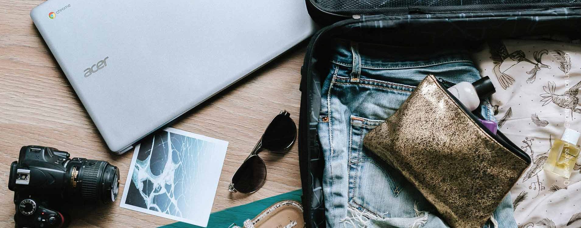 Utensilien für den Urlaub (Laptop, Koffer, Sonnenbrille) ausgebreitet auf einem Tisch, von oben fotografiert