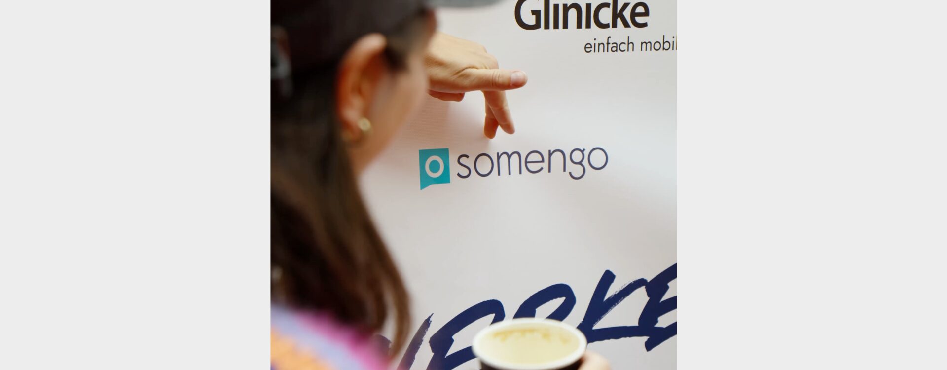 Frau zeigt mir ihren Fingern auf das Somengo-Logo einer Sponsoring-Wand